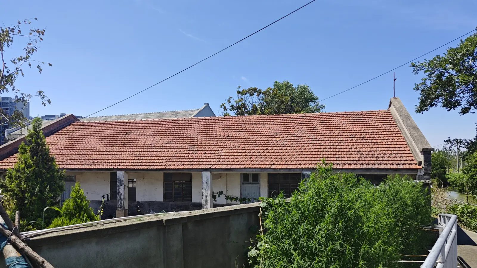 A long low building with a red tile roof and a small cross at the gable end behind a grey wall and green shrubs.