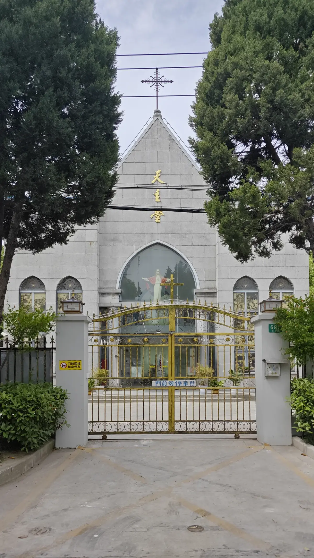 A grey gabled church front with gold Chinese characters behind a golden iron gate between pale gate posts, framed by trees.