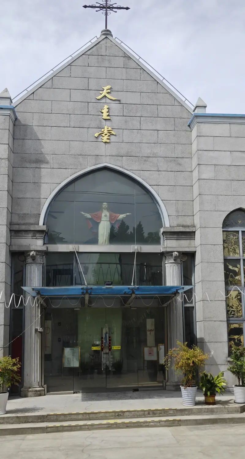 A grey stone church front with a pointed gable and cross, gold characters, a large arched glass window and a glazed entrance porch.