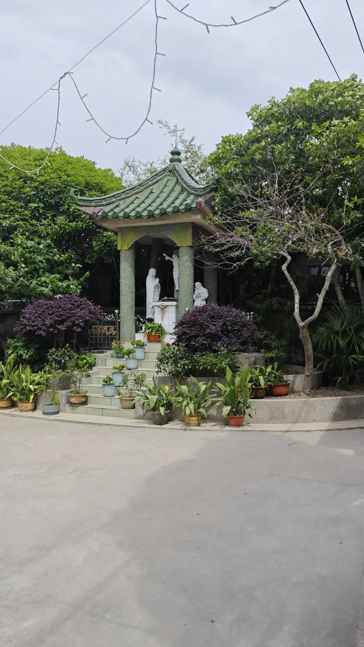 A small pavilion with a green tiled roof over a white statue, on a stepped base among flowering plants and trees.