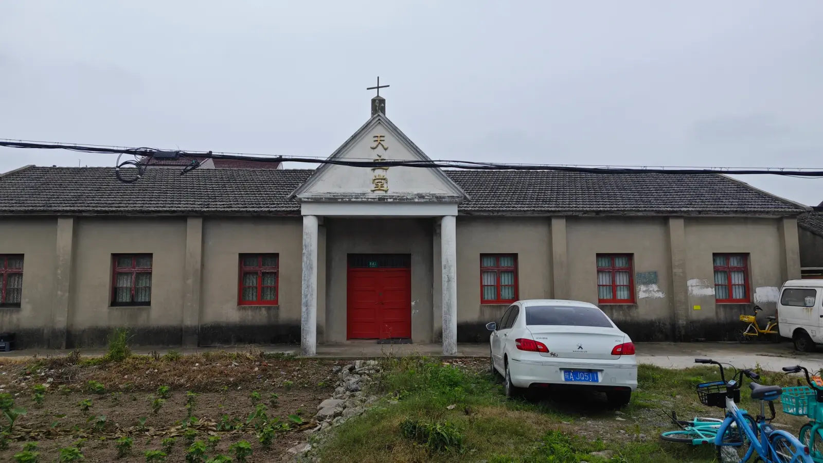 A single-storey grey building with a tiled roof, a white pedimented porch with a cross and Chinese characters, and red doors and window frames.