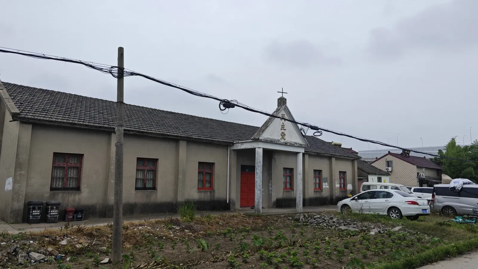 A long grey single-storey hall with red-framed windows and a white porch gable with a cross, behind a vegetable plot and a utility pole.