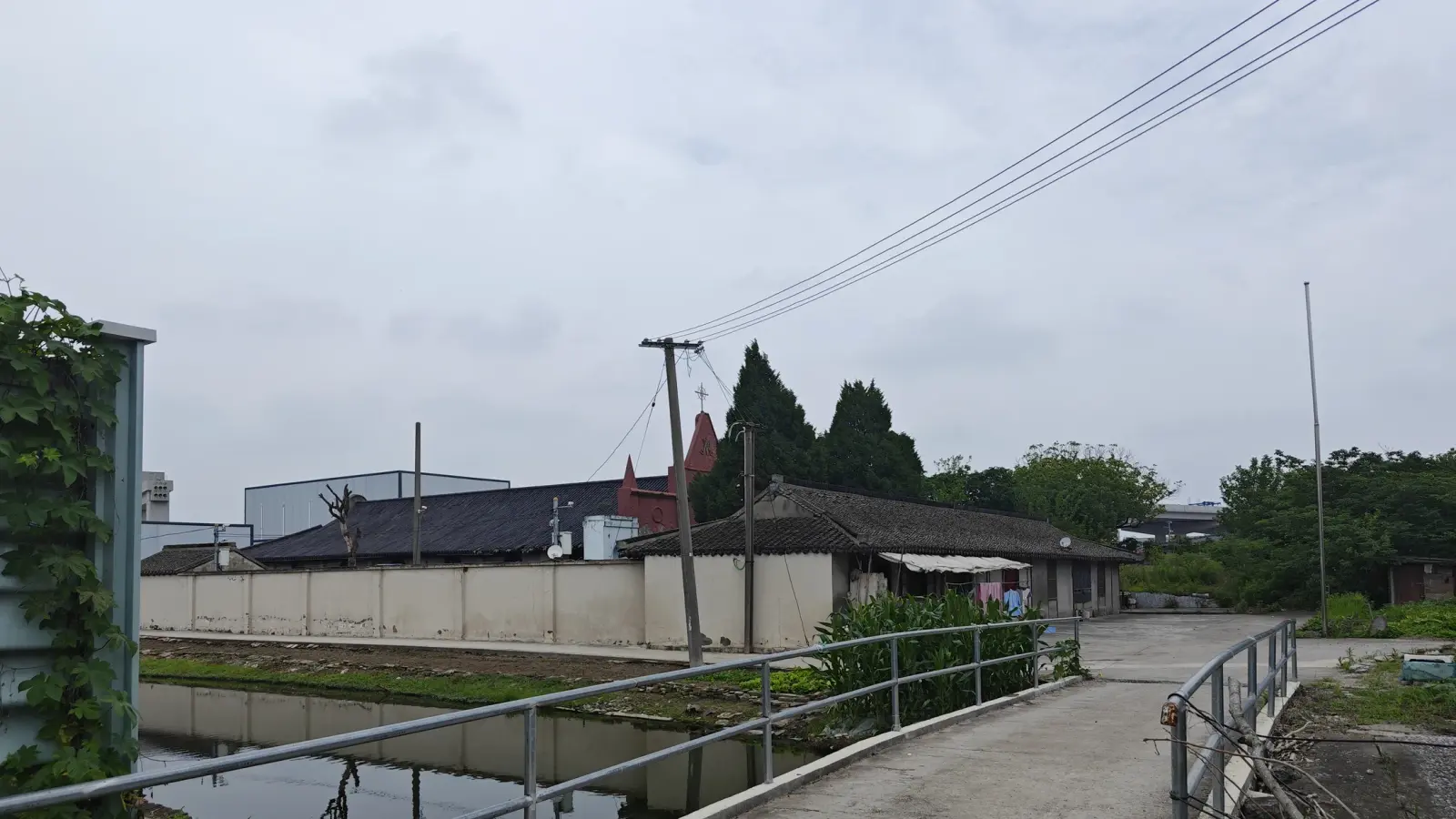 A footpath beside a small canal with a long wall and low tiled buildings, and a red church spire in the distance.