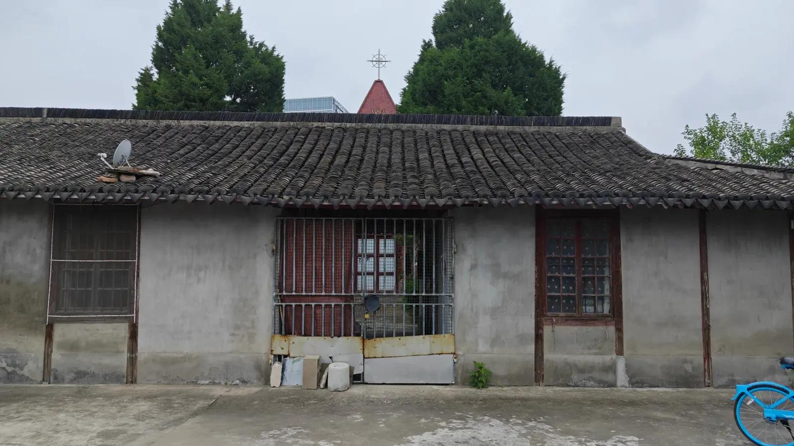 A grey single-storey building with a dark tile roof and barred windows, with a small red spire and cross behind it.