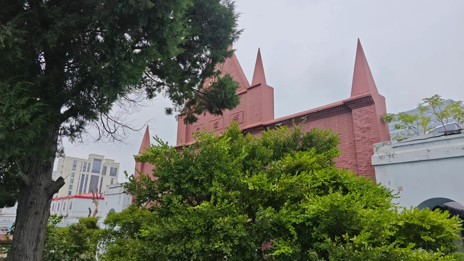 A red-pink church with several pointed spires above green foliage, with a pale modern building beyond.