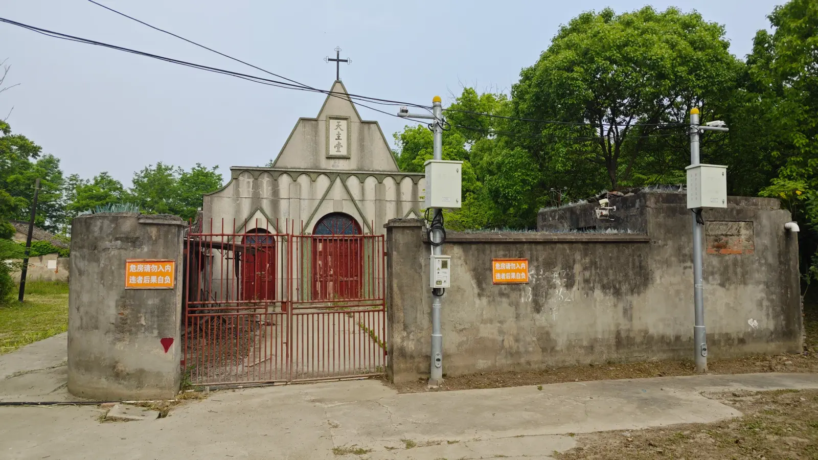 A small grey church front with a pointed gable and cross behind a red iron gate set in a stained concrete wall, with trees behind.