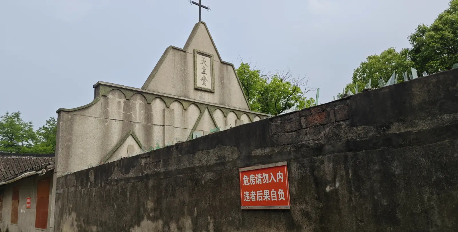 A grey gable with a cross and a small name board above a dark brick wall with a red notice.