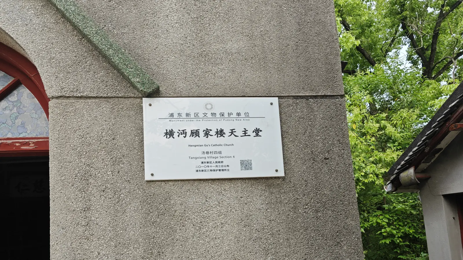 A white plaque with Chinese and English text reading Monument under the Protection of Pudong New Area, fixed to a pebbledash wall.