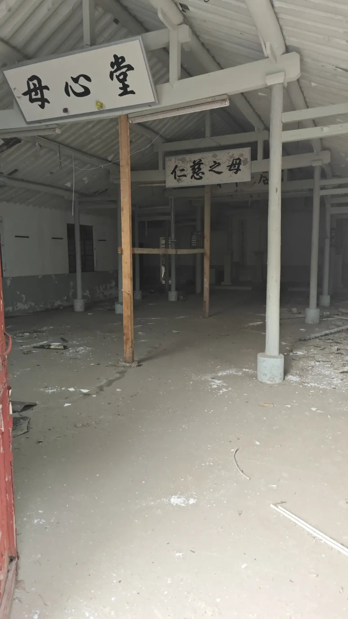 A bare, dusty hall with timber posts and two painted board signs in Chinese hanging from the roof frame.