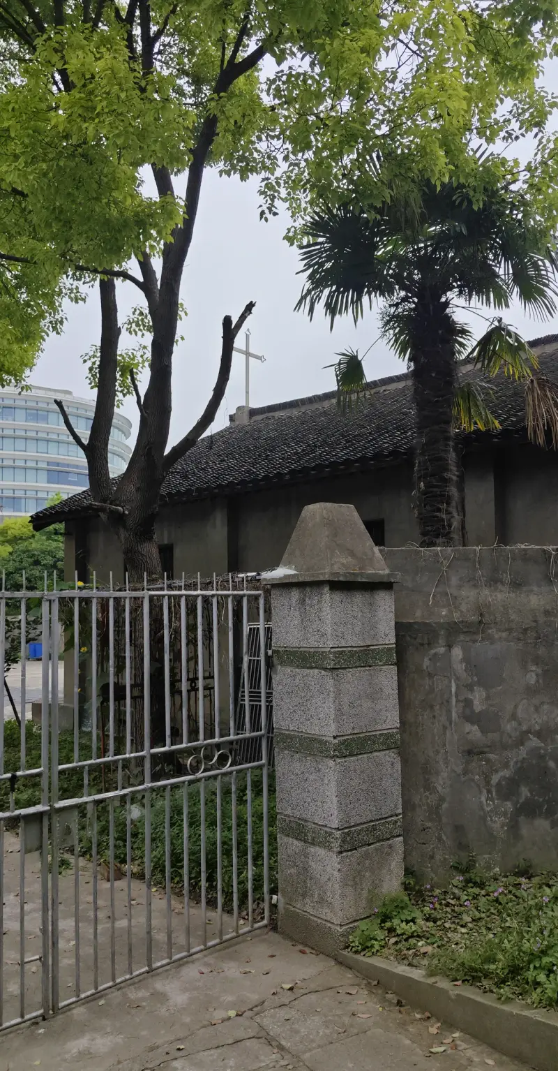 An iron gate with stone gate post beside a grey wall, a tiled roof and palm tree, with a glass office building beyond.