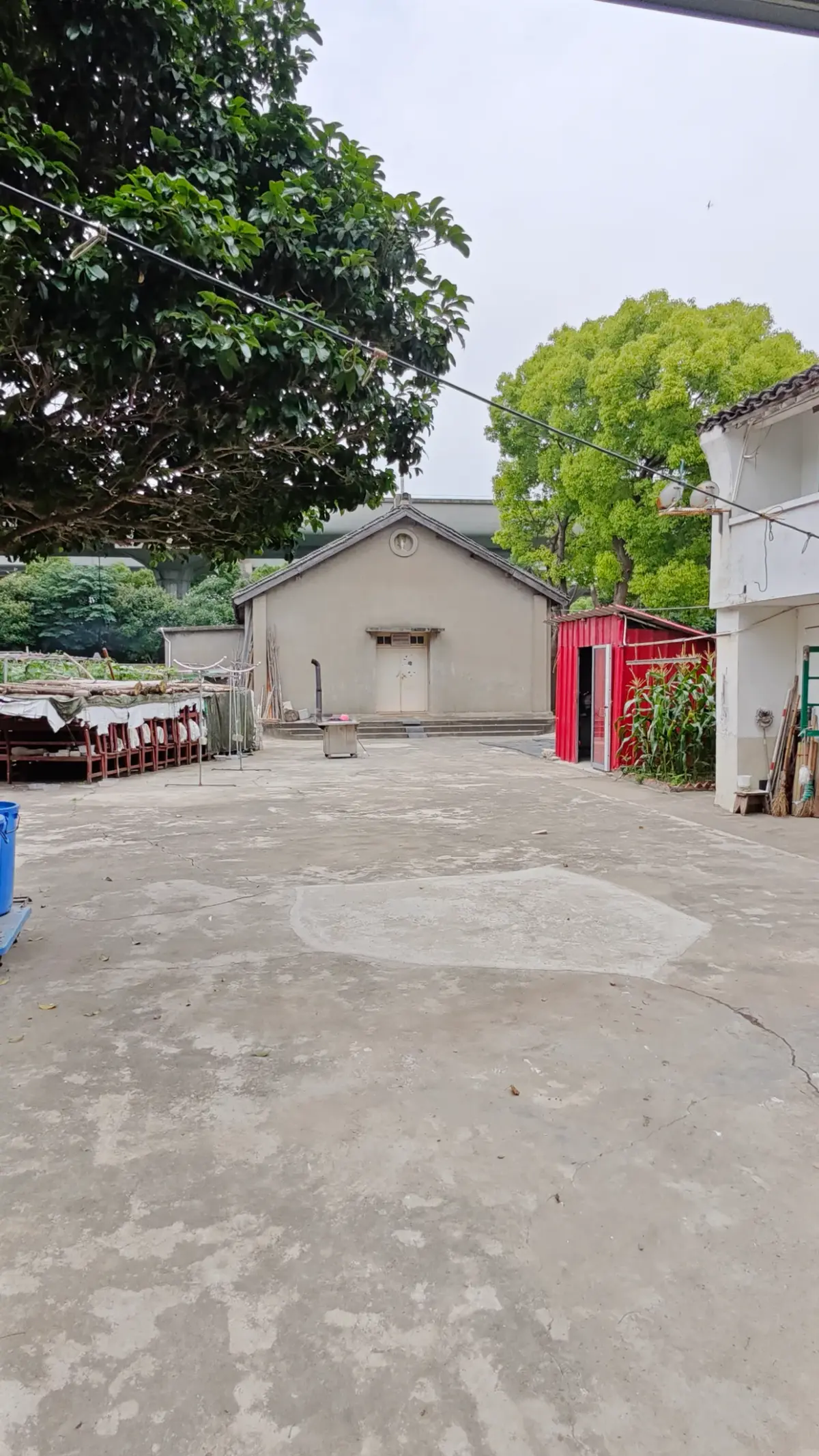 A wide concrete yard with a small grey gabled building at the back, a red container and trees.