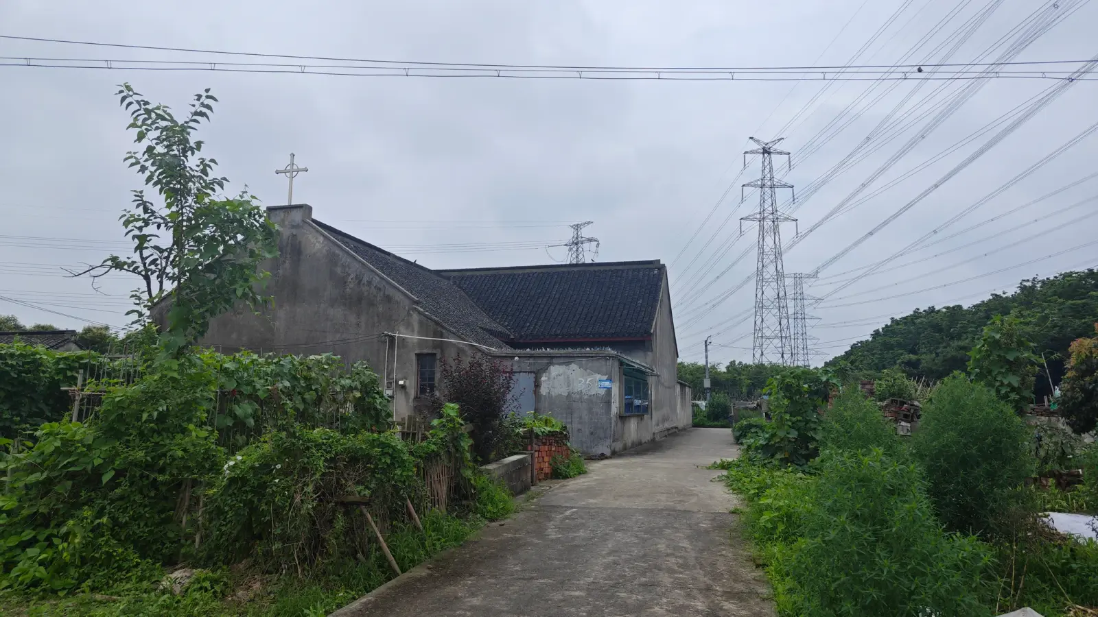 A grey church with a cross at its gable end beside a concrete lane lined with greenery and overhead power lines.