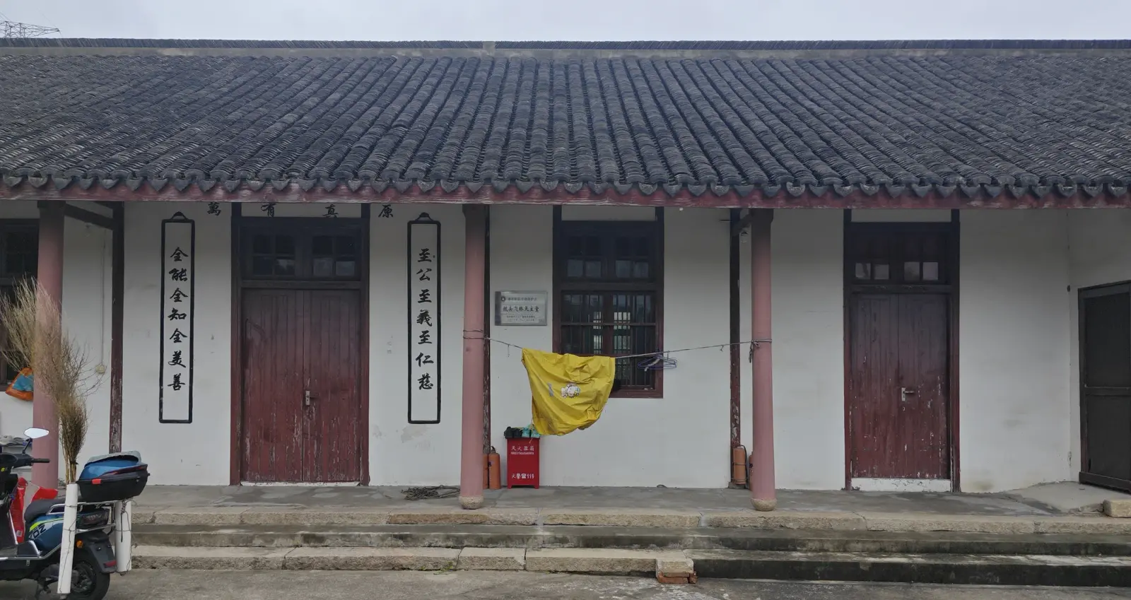 A whitewashed building front with a tiled roof, red pillars, dark red doors and vertical couplet boards.