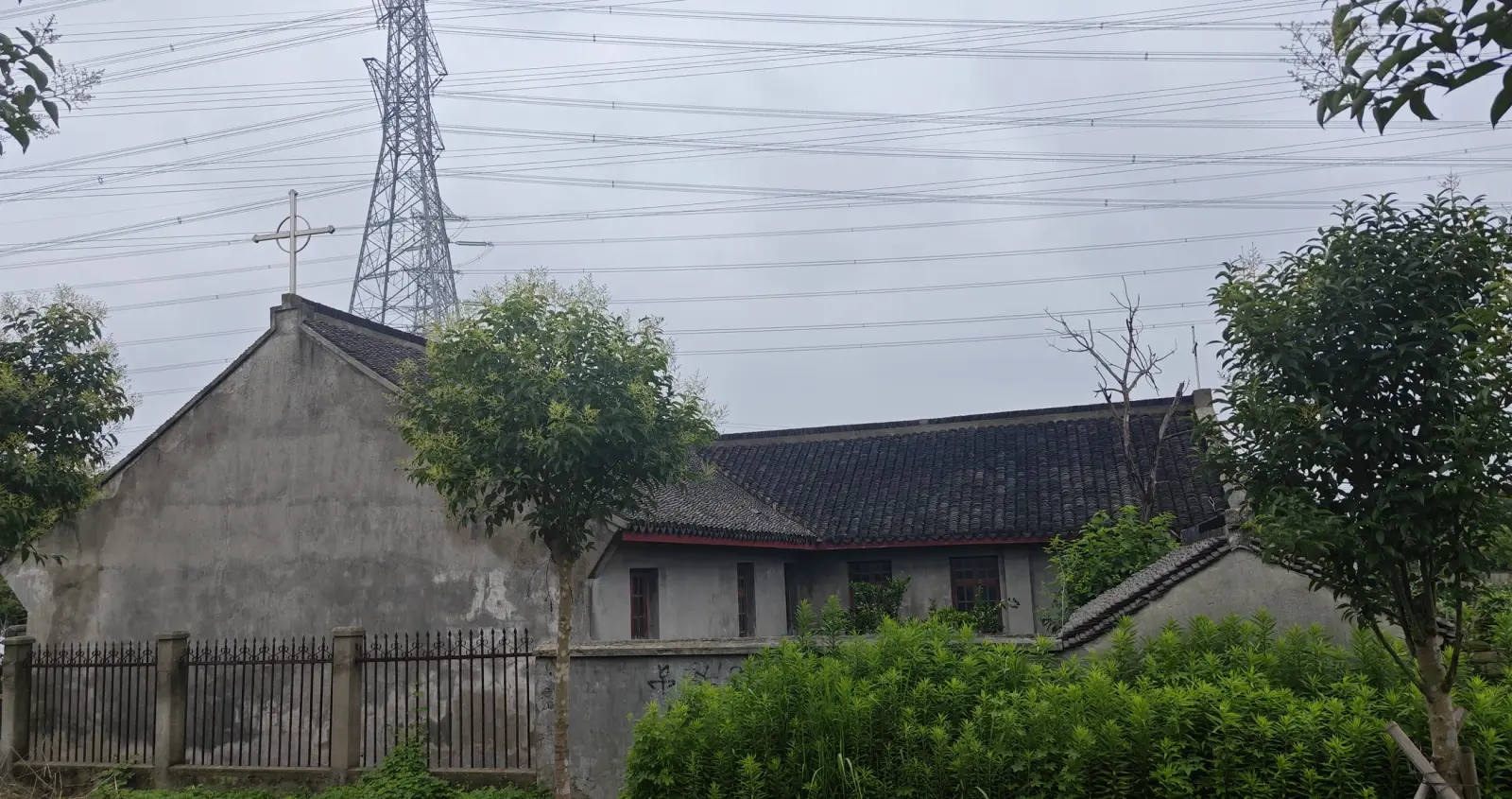 A grey church gable topped with a cross behind a fence and trees, with an electricity pylon in the background.