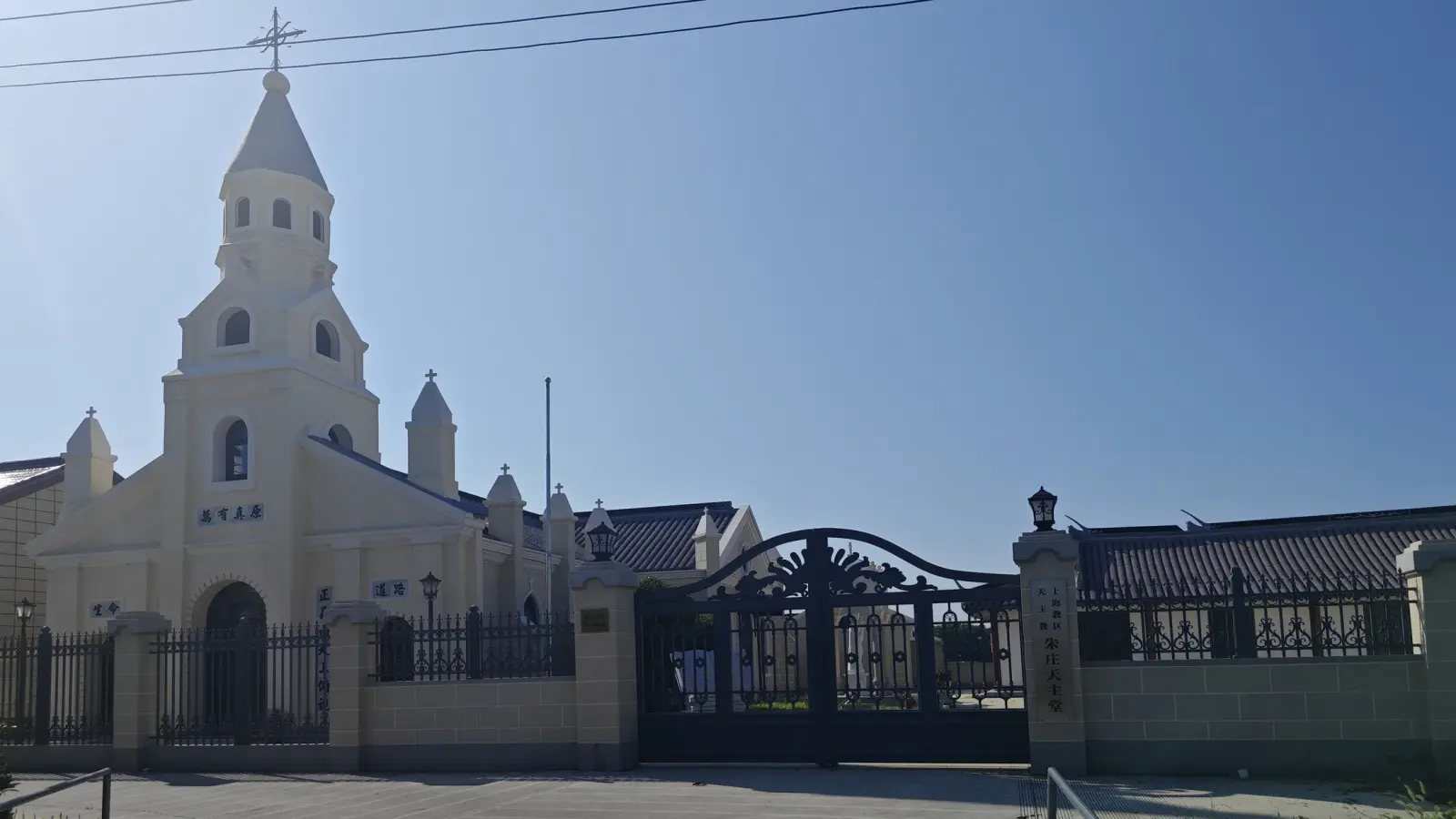 A cream-coloured church with a steeple and pinnacles behind a wall and a decorative iron gate under a clear blue sky.