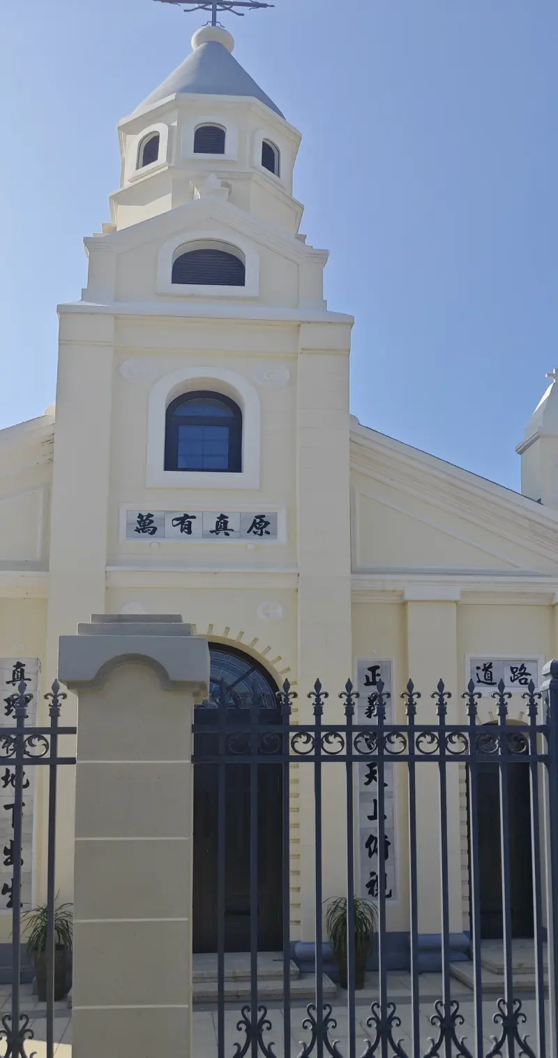 Cream church gable with a bell steeple and a board with four Chinese characters, seen through railings.