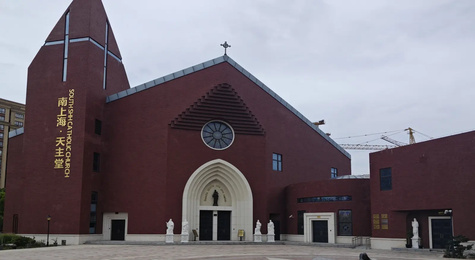 A red-brick modern church with a tall sloping tower carrying a cross, a round window and a pointed arched entrance.