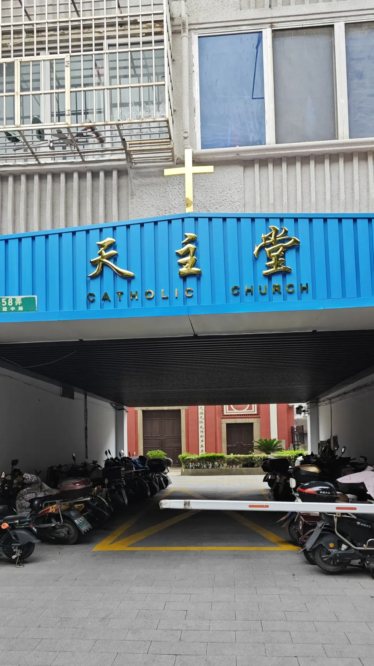 Blue sign over a passageway reading the Chinese characters for Catholic church and CATHOLIC CHURCH, with the church visible at the far end.