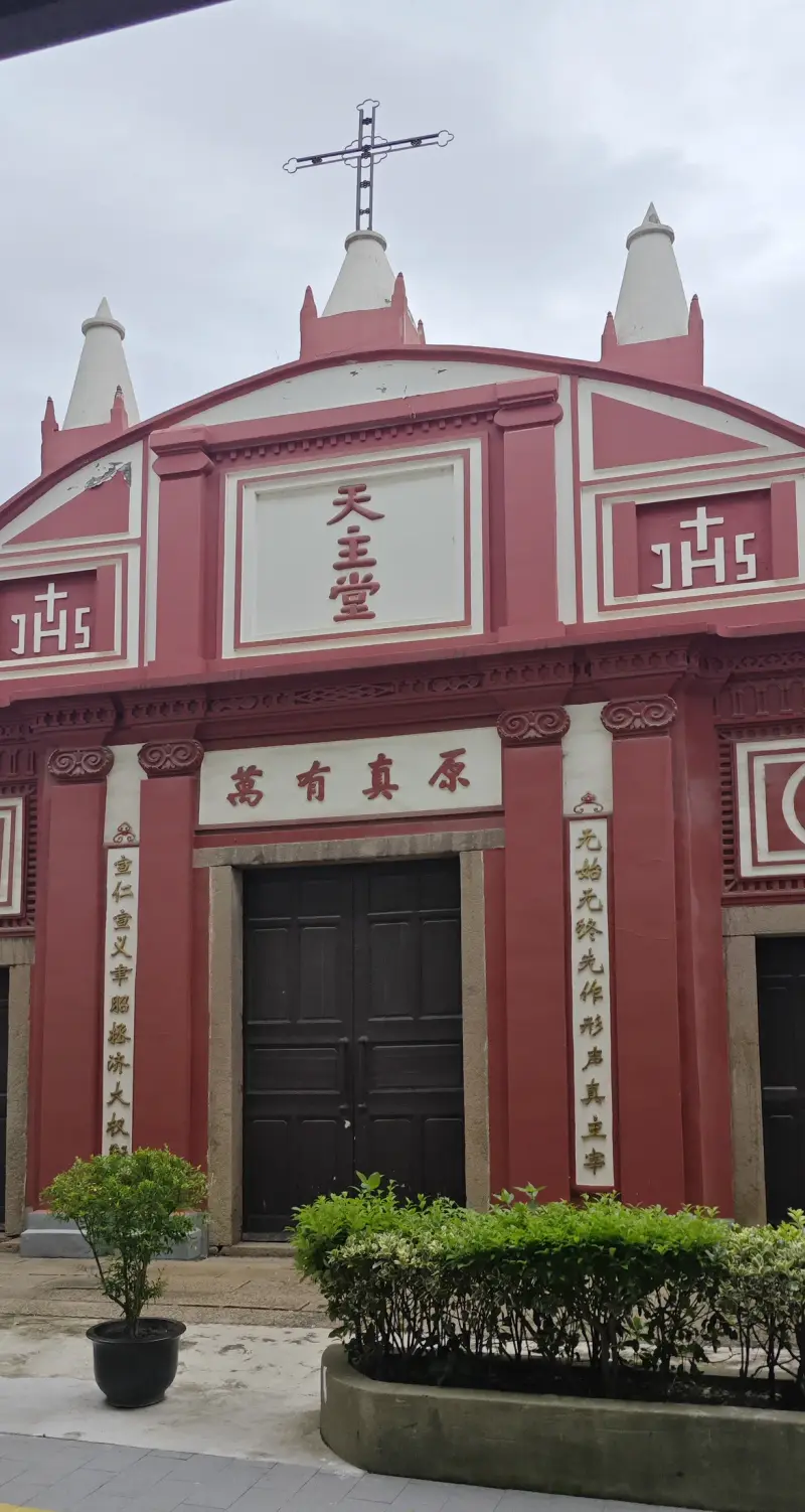 Red and white church facade with a curved gable, two small pinnacles, a cross and the characters for Catholic church.