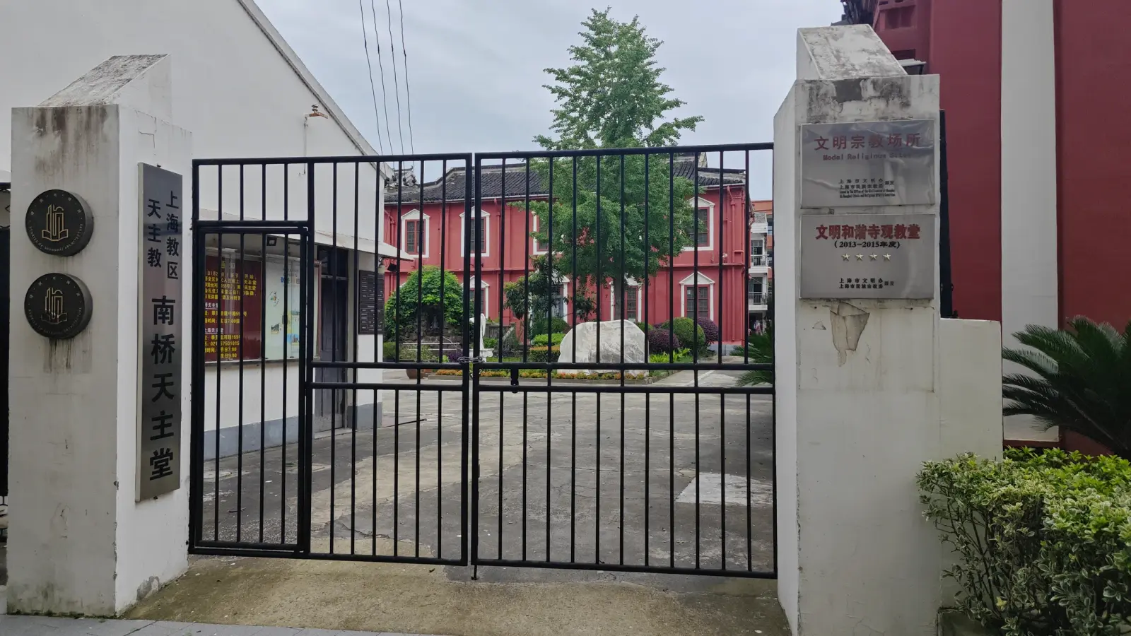 A red church building across a courtyard, viewed through a black iron gate between white gateposts.