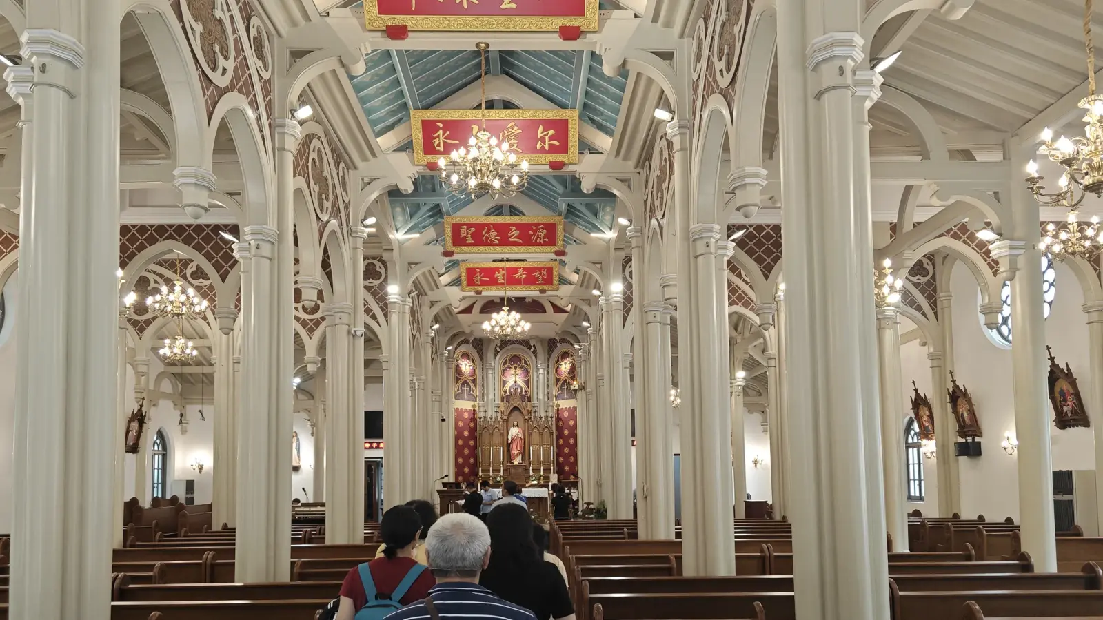 A church interior with tall cream columns, a decorated ceiling and red banners with gold characters above wooden pews.
