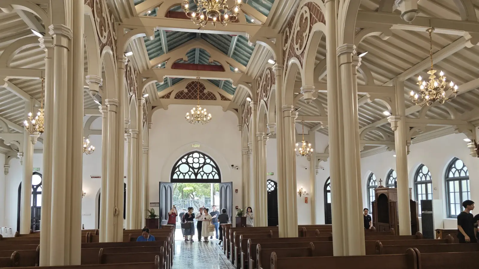 A long nave with pale columns and wooden pews leading to an open door with a pointed arched window.