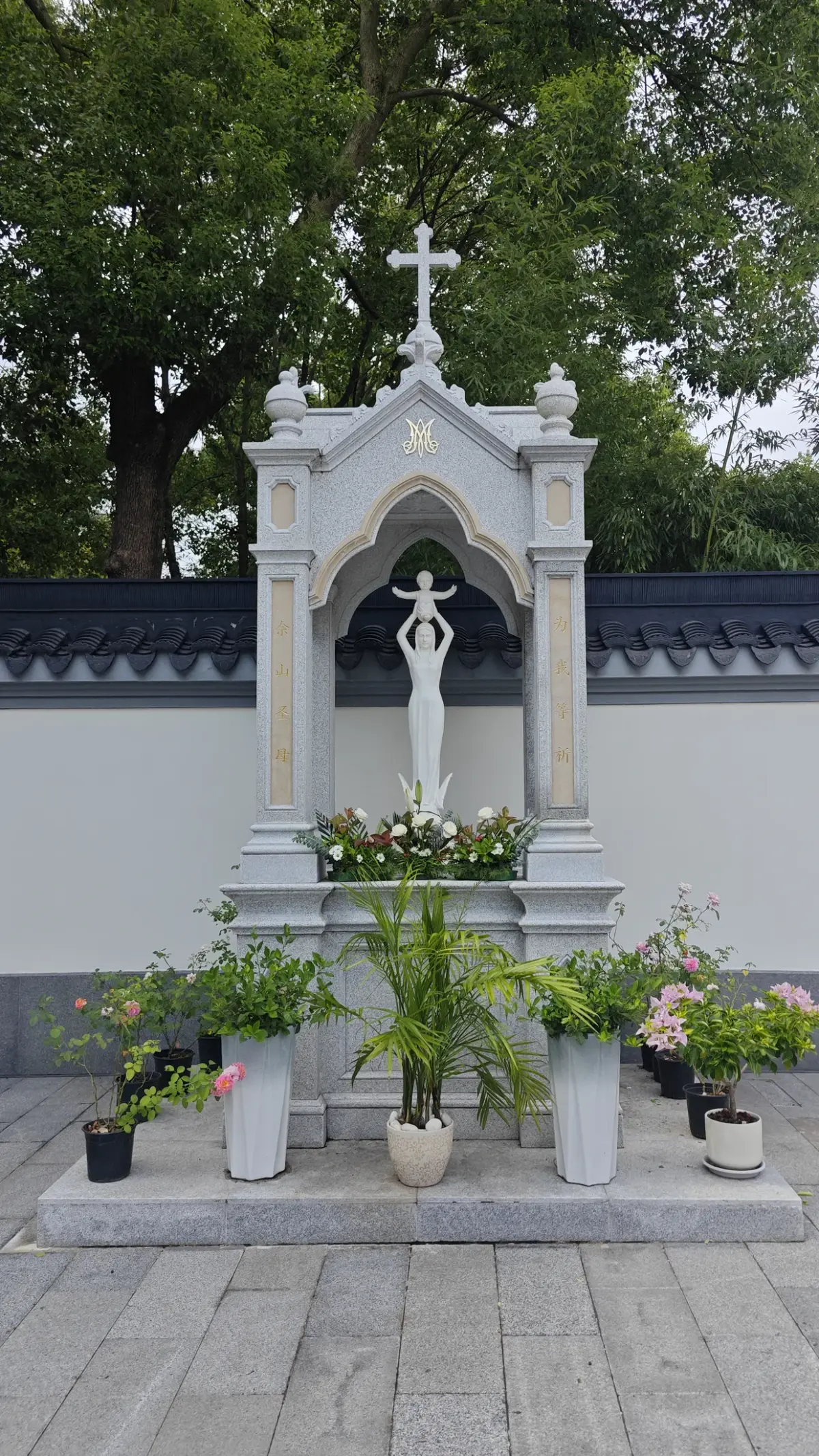 A white statue under a pointed stone canopy topped with a cross, with flowers and plants at its base.