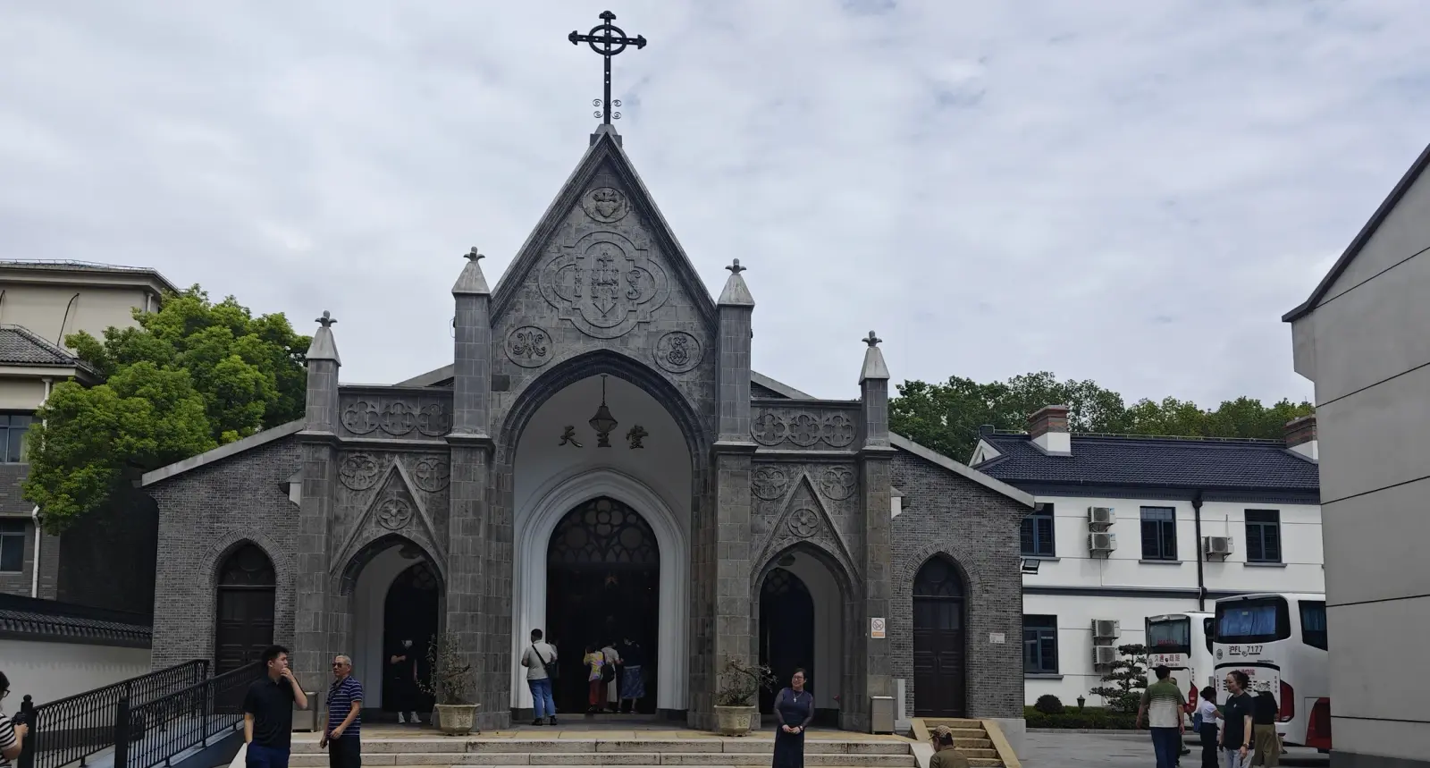 A grey stone church front with a tall pointed gable, carved tracery, a cross at the top and visitors on the steps.