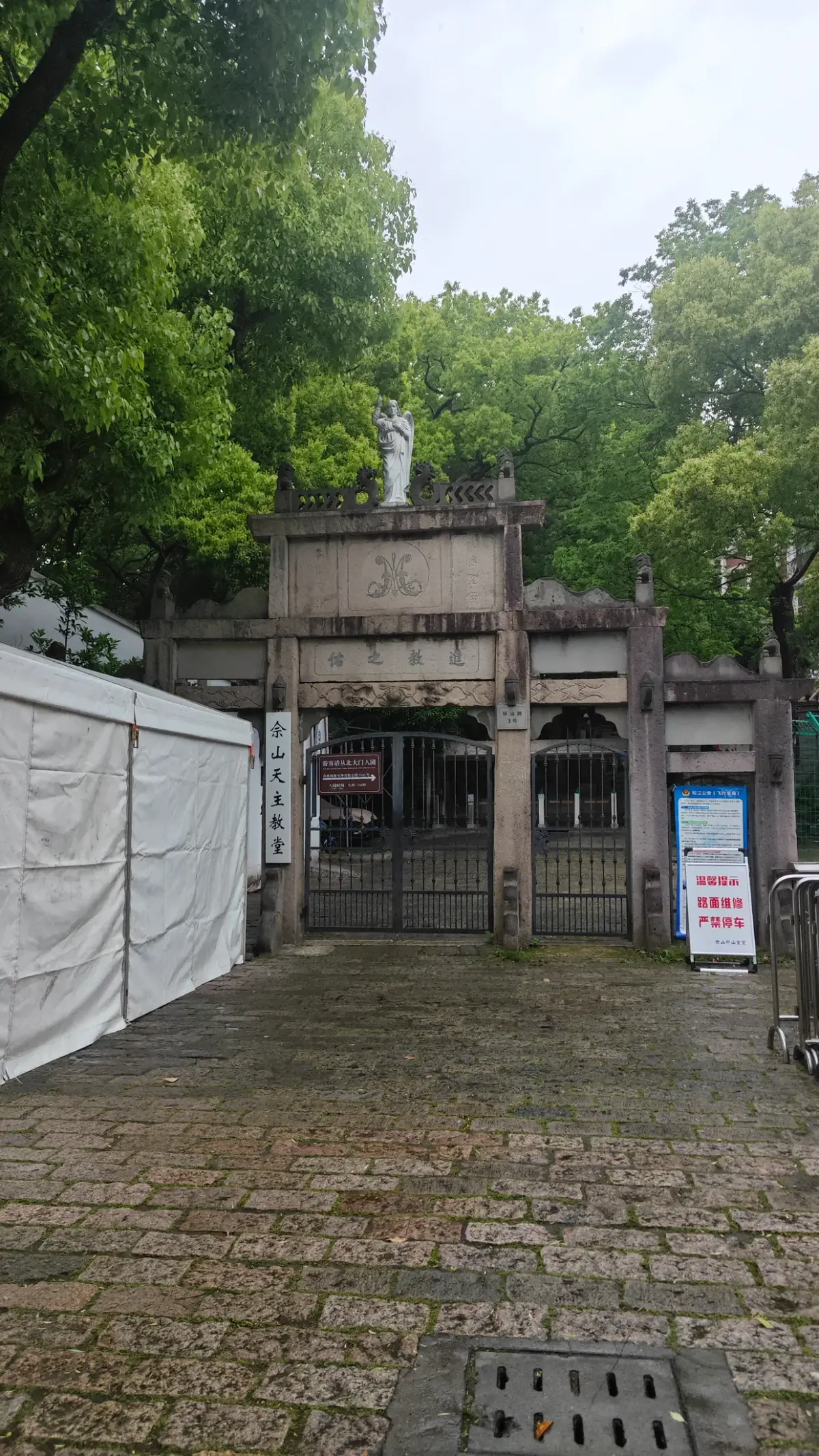 An old stone gateway with iron gates at the end of a paved path among trees.