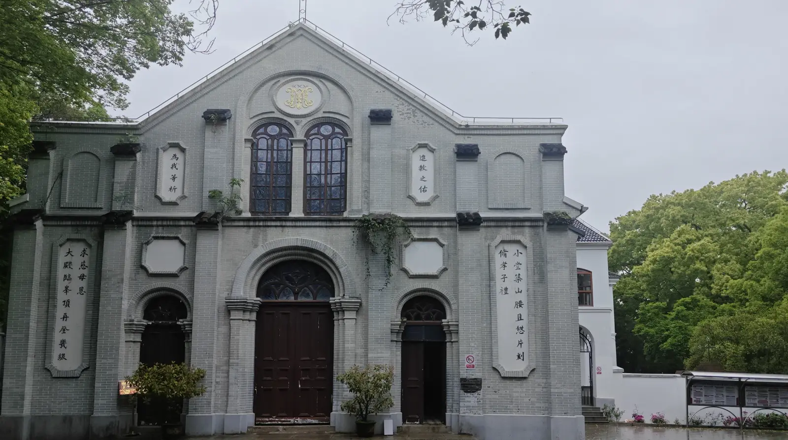 A grey stone church front with a gabled top, two tall arched windows, vertical inscribed panels and arched wooden doors, framed by trees.