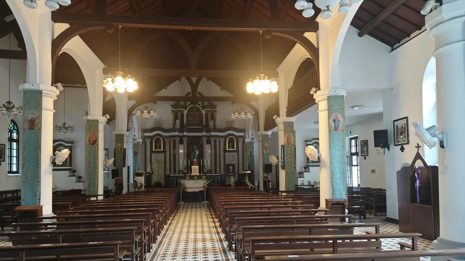 A church interior with a chequered tile aisle, rows of pews, turquoise columns and a timber roof.