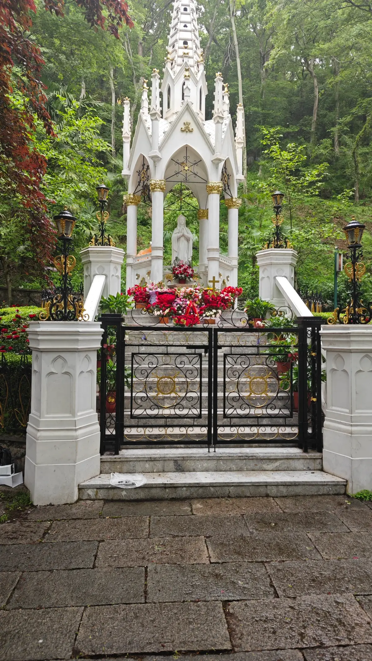 A white open-sided shrine with a pointed canopy above steps and an iron gate, surrounded by green trees.
