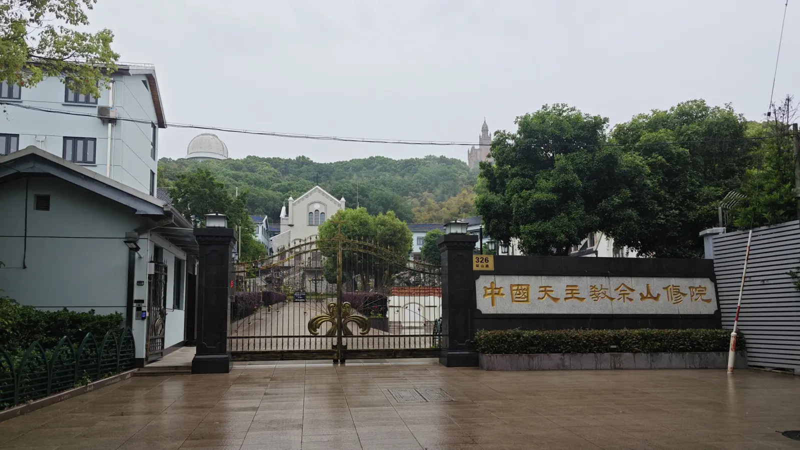A black iron gate between stone pillars, with a long sign in gold Chinese characters and wooded hill behind.