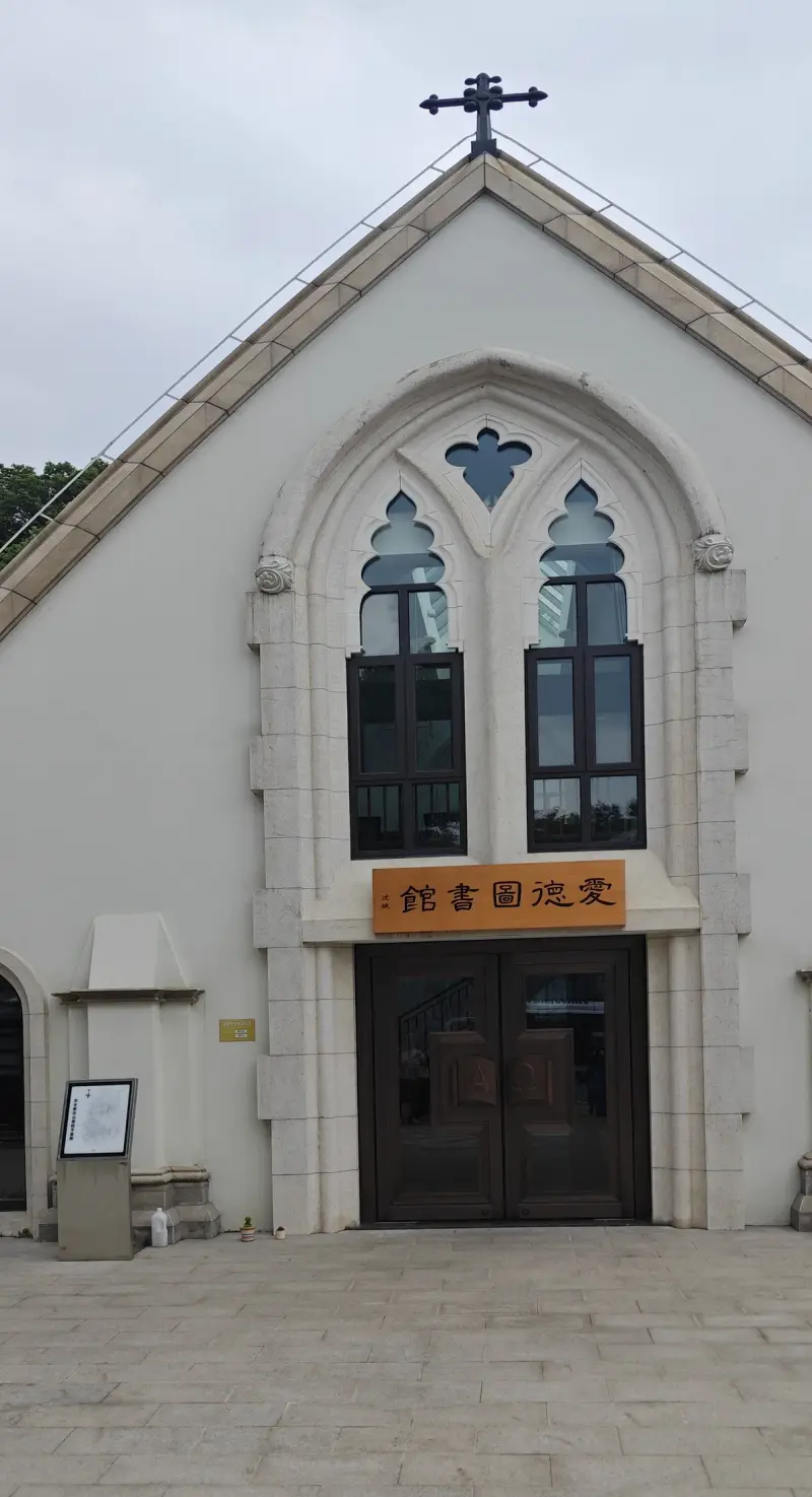 A pale chapel front with a steep gable, a tall pointed window and a cross at the peak, above a wooden double door.