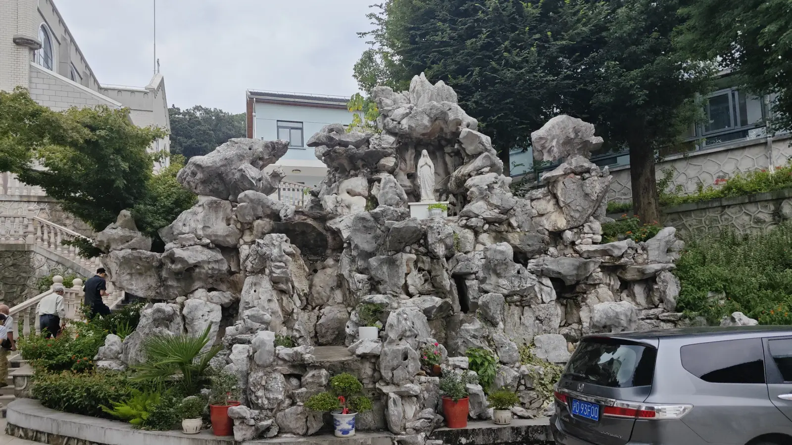 A tall pile of pale rocks with a statue in a niche, ringed by potted plants beside a car.