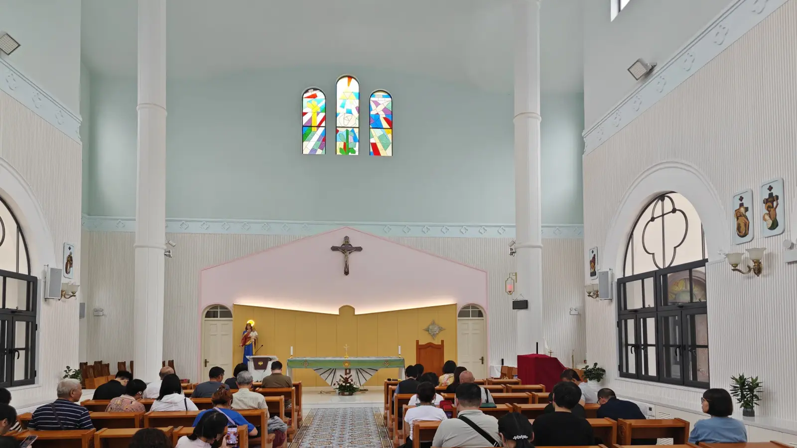 The chapel interior with pale blue walls, worshippers on wooden pews and a stained glass window above the altar.