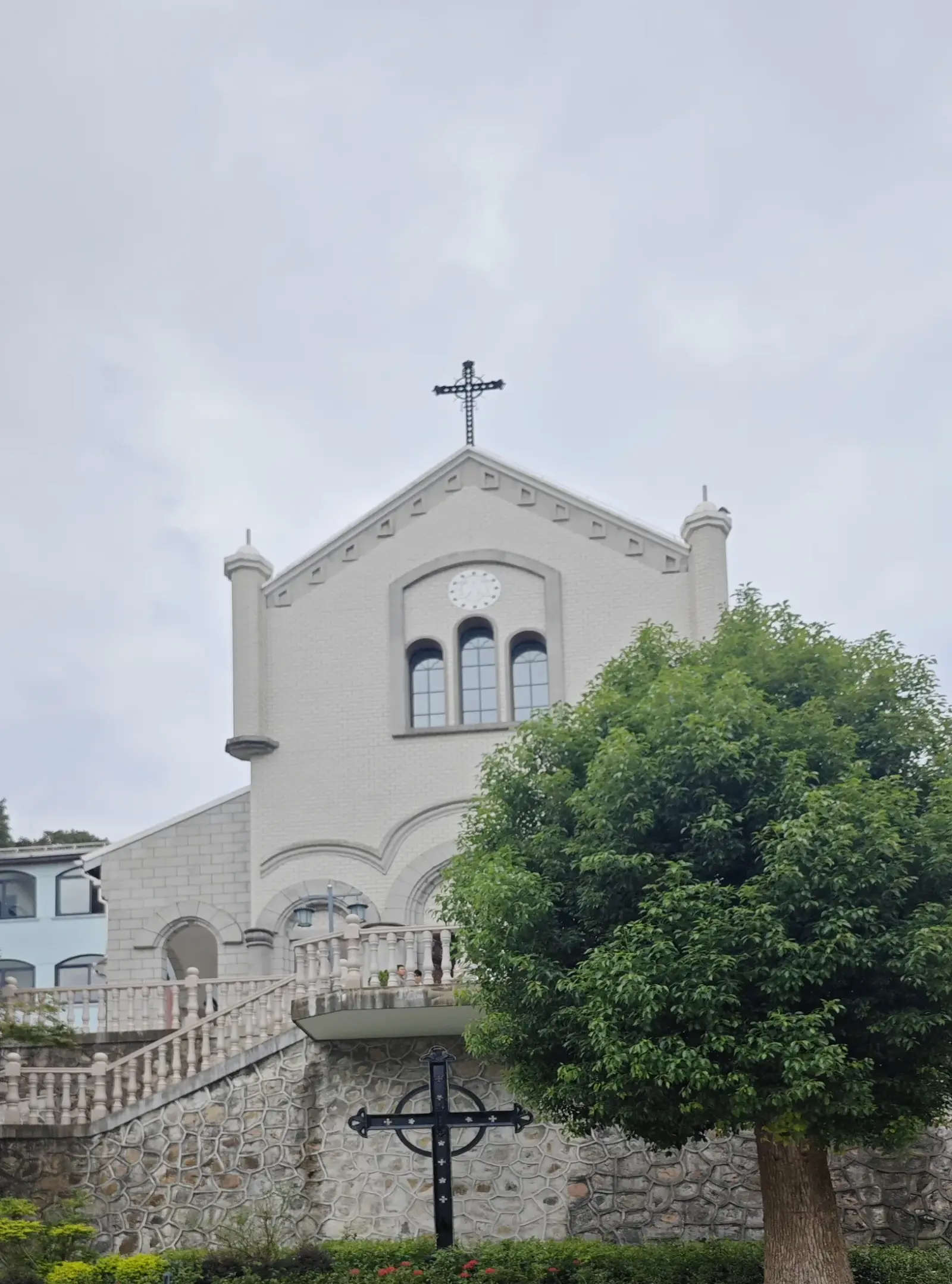 A pale chapel with a cross on its gable above a stone wall and a green tree.