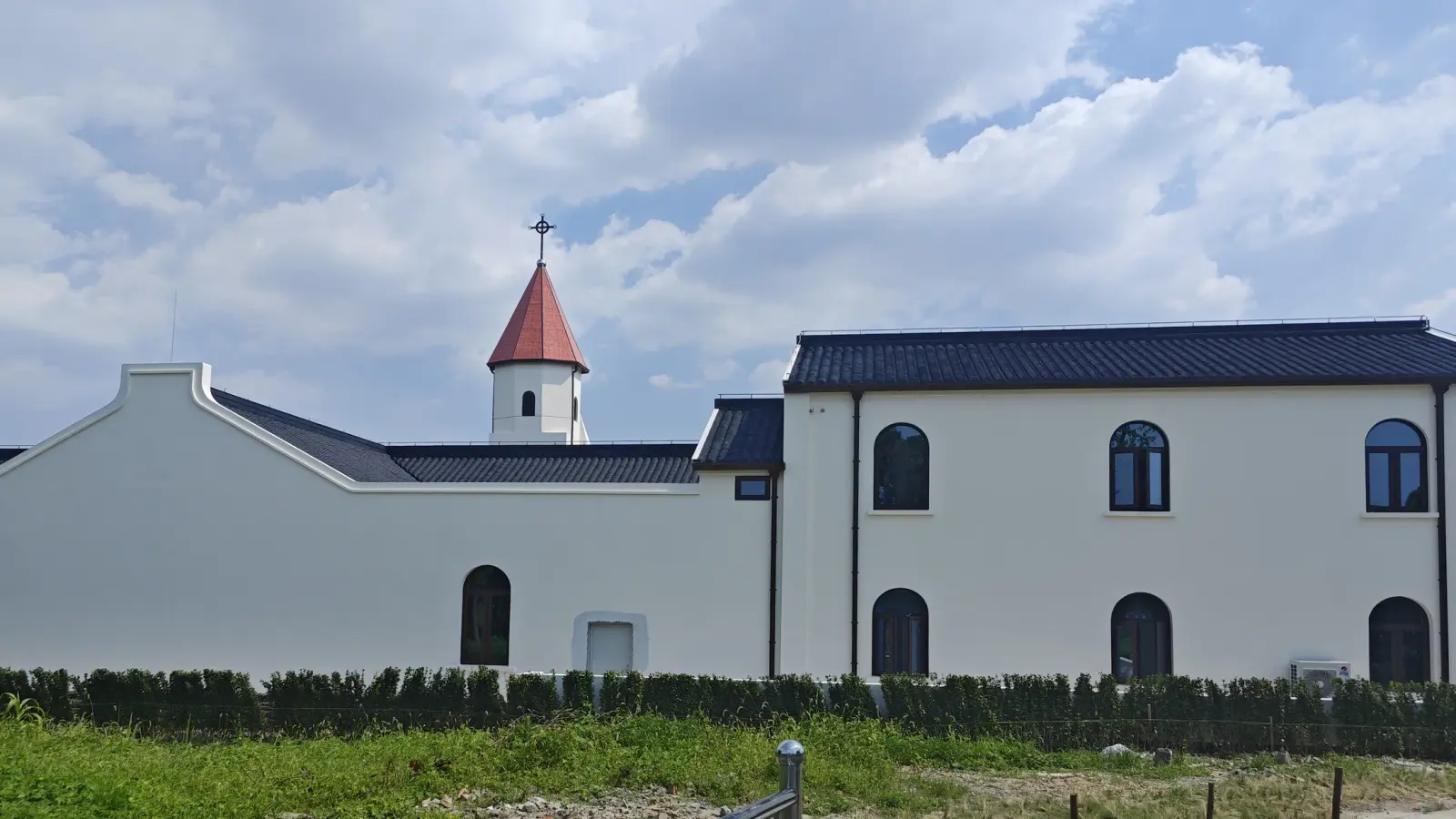 A long white wall with dark arched windows and a red-roofed tower rising behind it, beyond a grass verge.