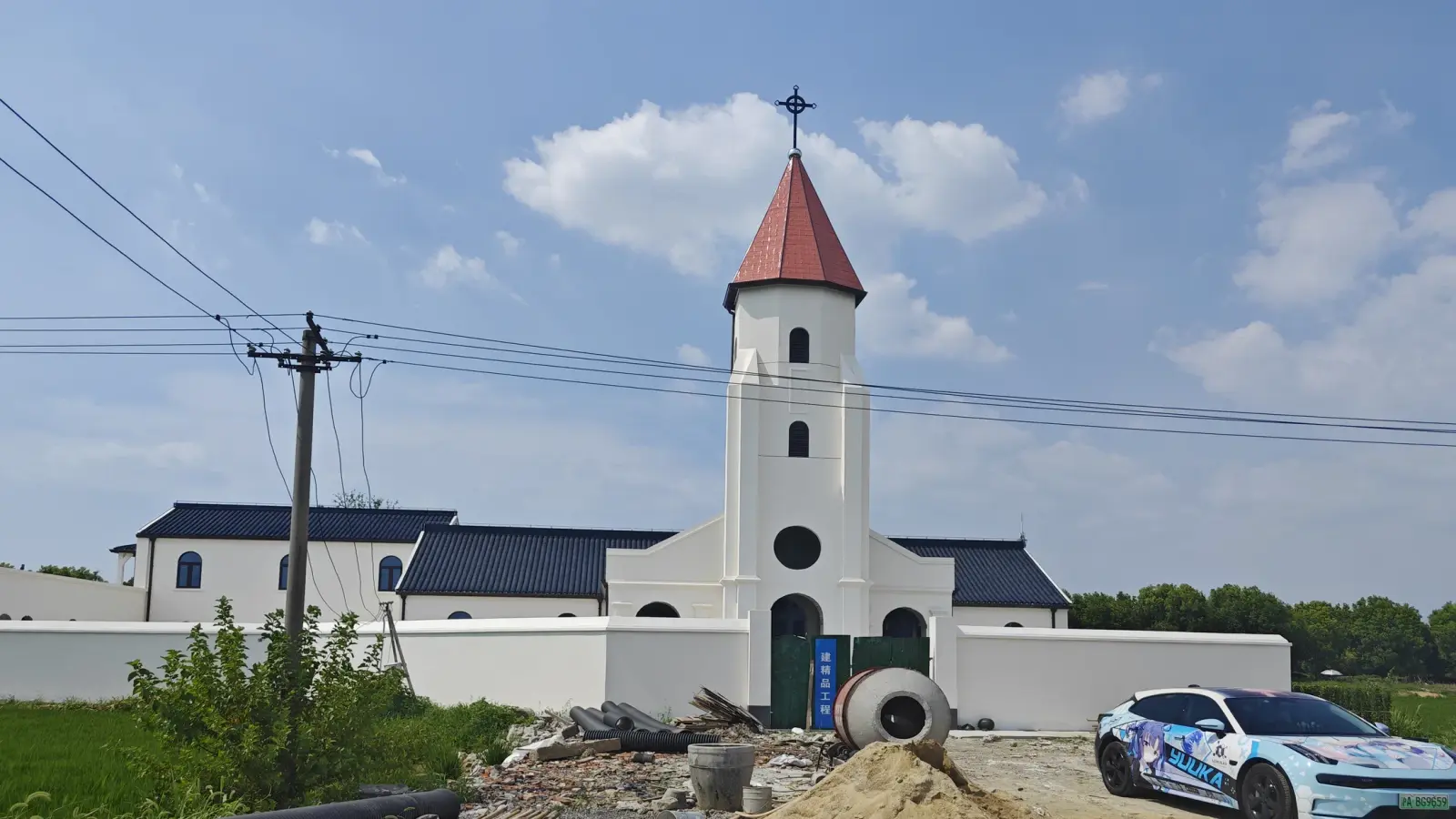 A white tower with a red pointed roof and cross above low dark-tiled roofs and a white wall.