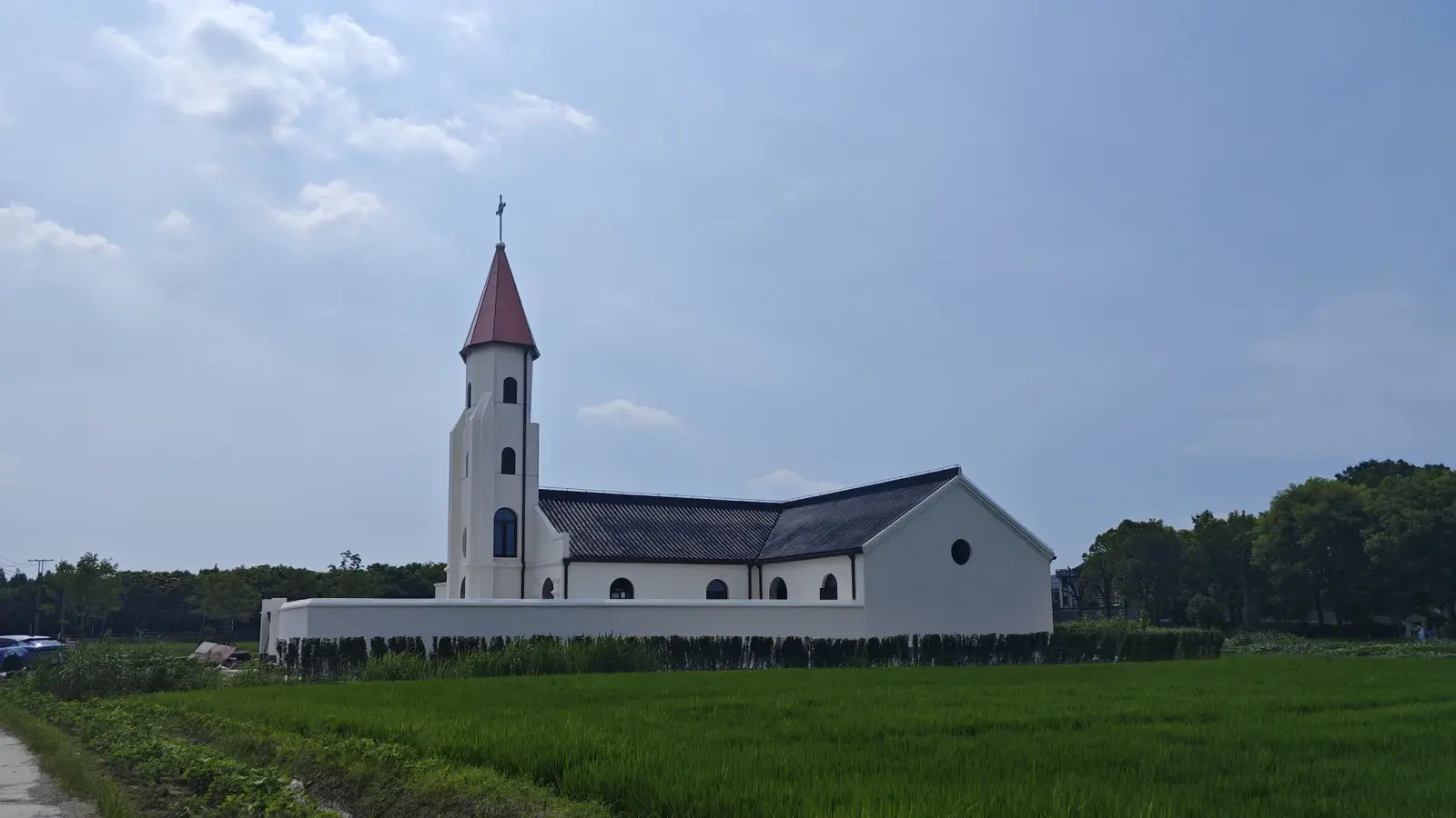 A white church with a slim tower topped by a red pointed roof stands behind a wall at the far side of a green field.