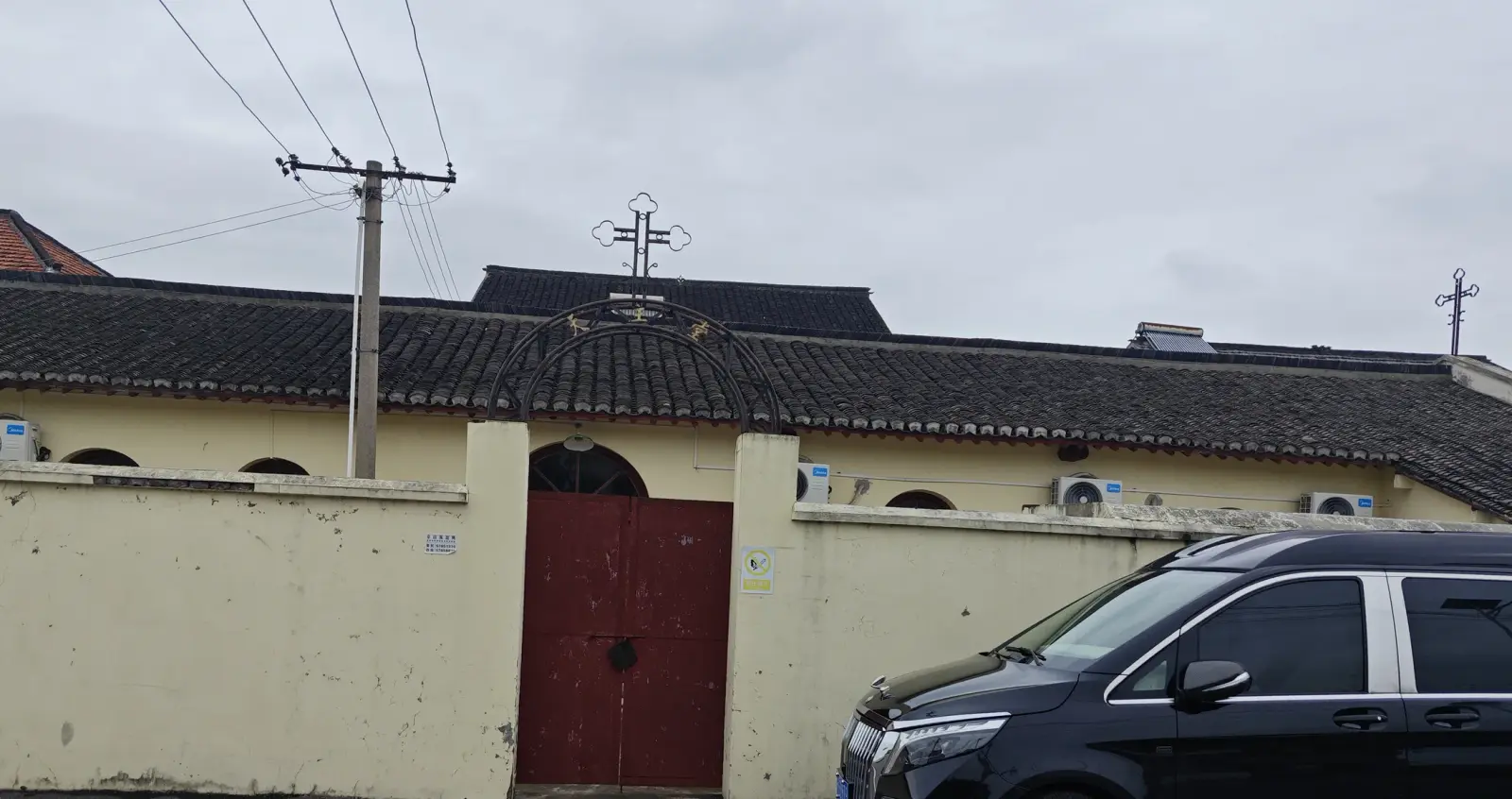 A cream-coloured wall with a red arched gate, a grey-tiled roof and an iron cross above it, with a black car parked in front.