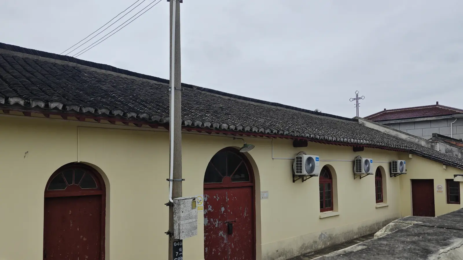 A long single-storey cream wall with two arched red doors and several air-conditioning units under a dark tiled roof, with a cross on the ridge.
