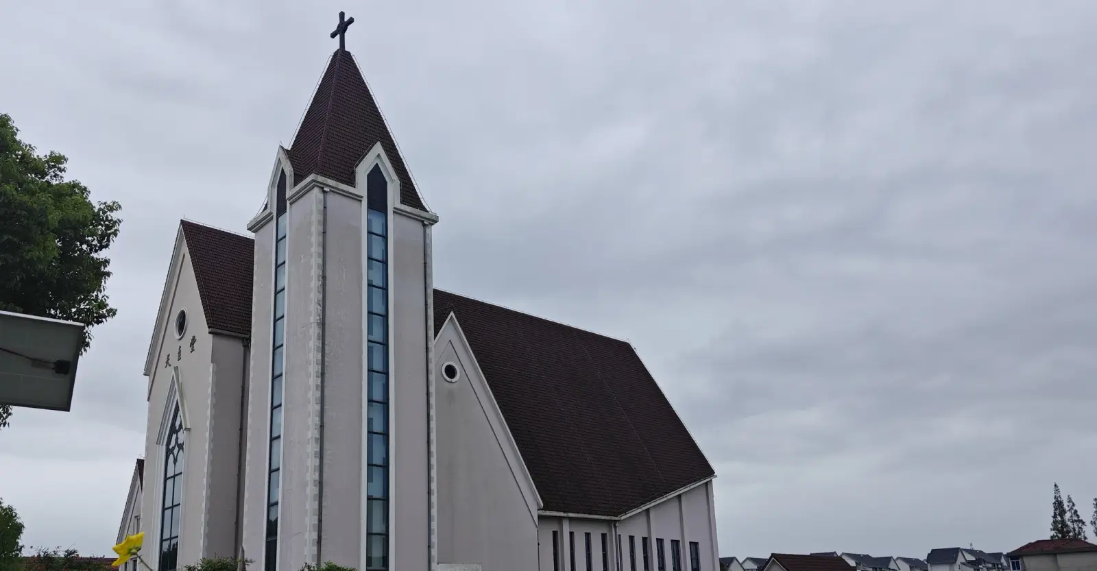 A tall cream tower with a dark pointed roof and thin blue window next to a dark gabled roof, against a grey sky.