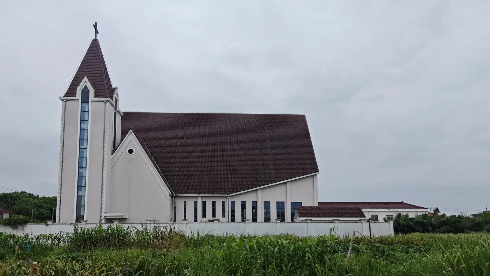 A pale cream church with a tall narrow tower with a cross and a long glazed window, beside a steep dark red roof, across a field.