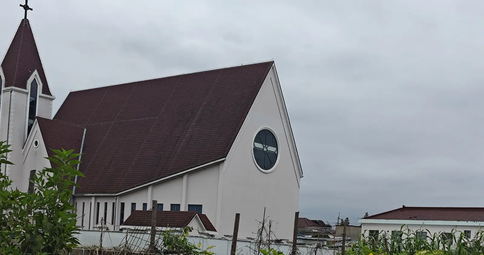 A cream church with a steep dark red roof, a round window in the gable and a tower at left, beyond a field.