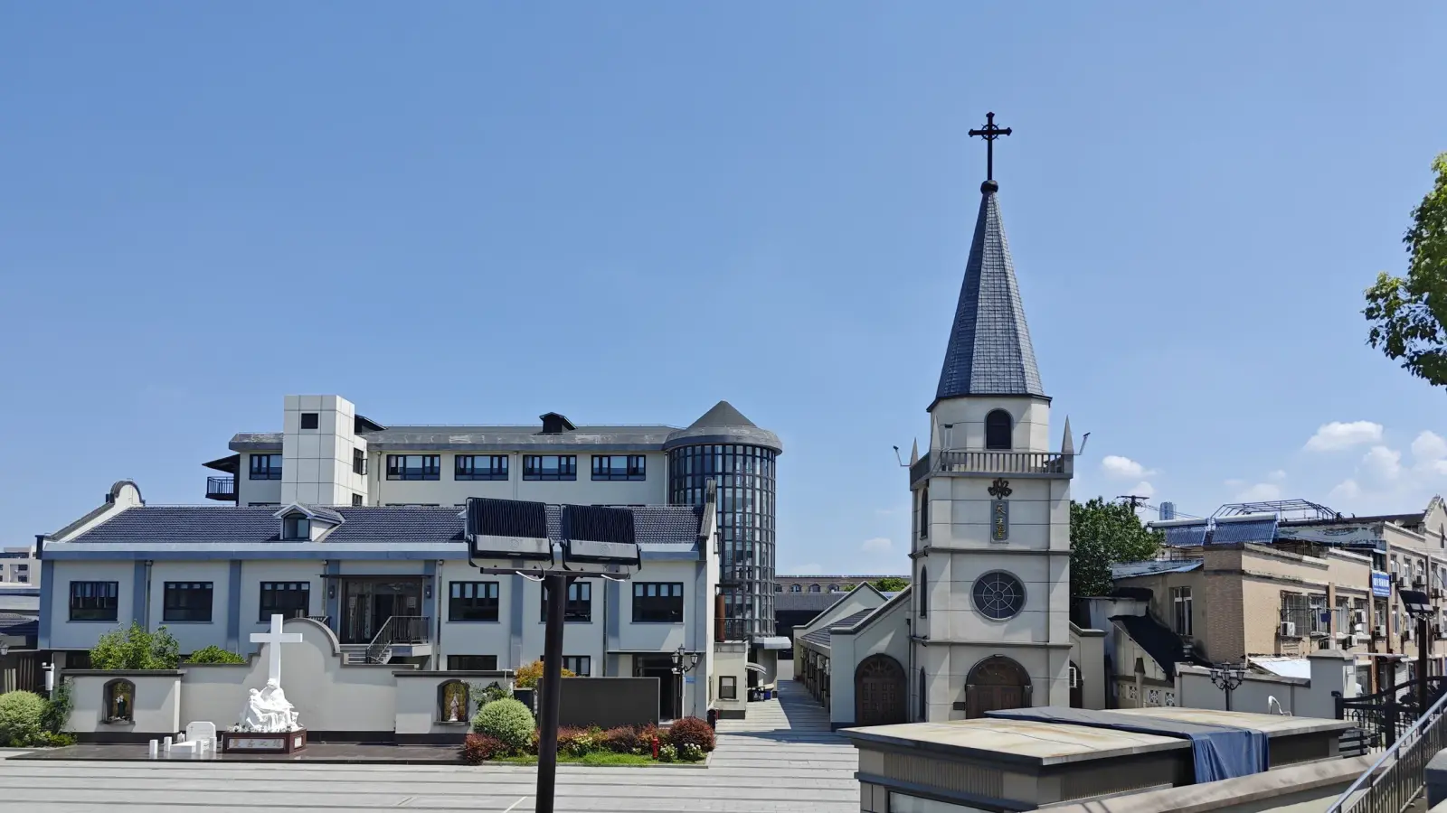 A courtyard view with a white bell tower beside a low grey-roofed church building and a taller modern block behind.