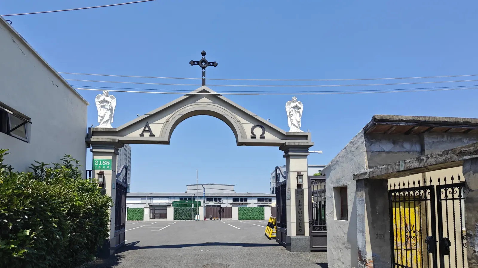 A stone arch marked with Alpha and Omega, crowned by a cross and two white angel statues, opening onto a courtyard.