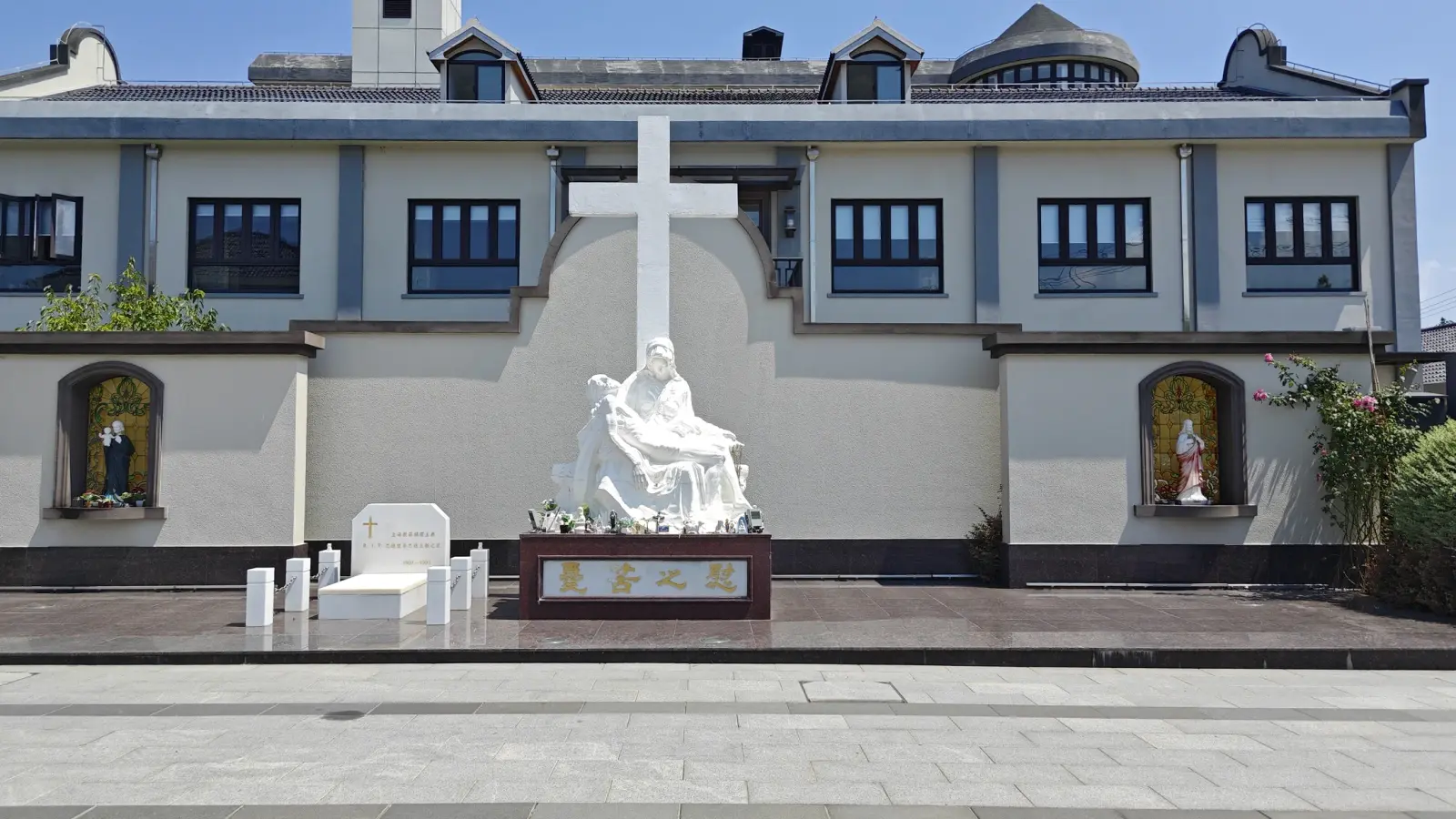 A white sculpture of Mary holding Christ beneath a large white cross on a cream wall with two arched shrines.