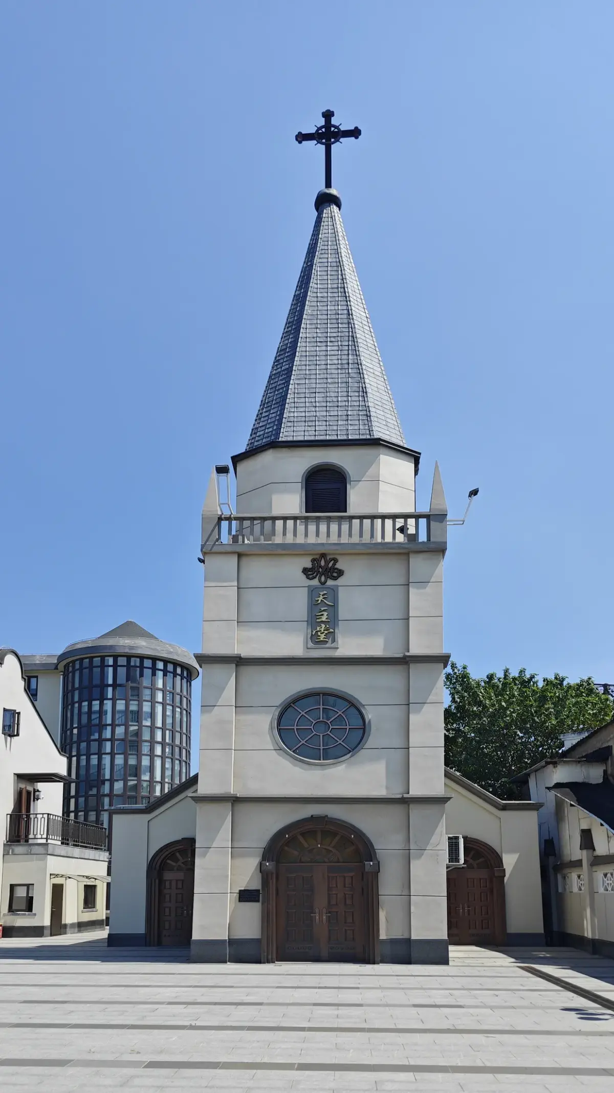 A white church bell tower with a pointed spire and cross rising above a paved courtyard under a blue sky.
