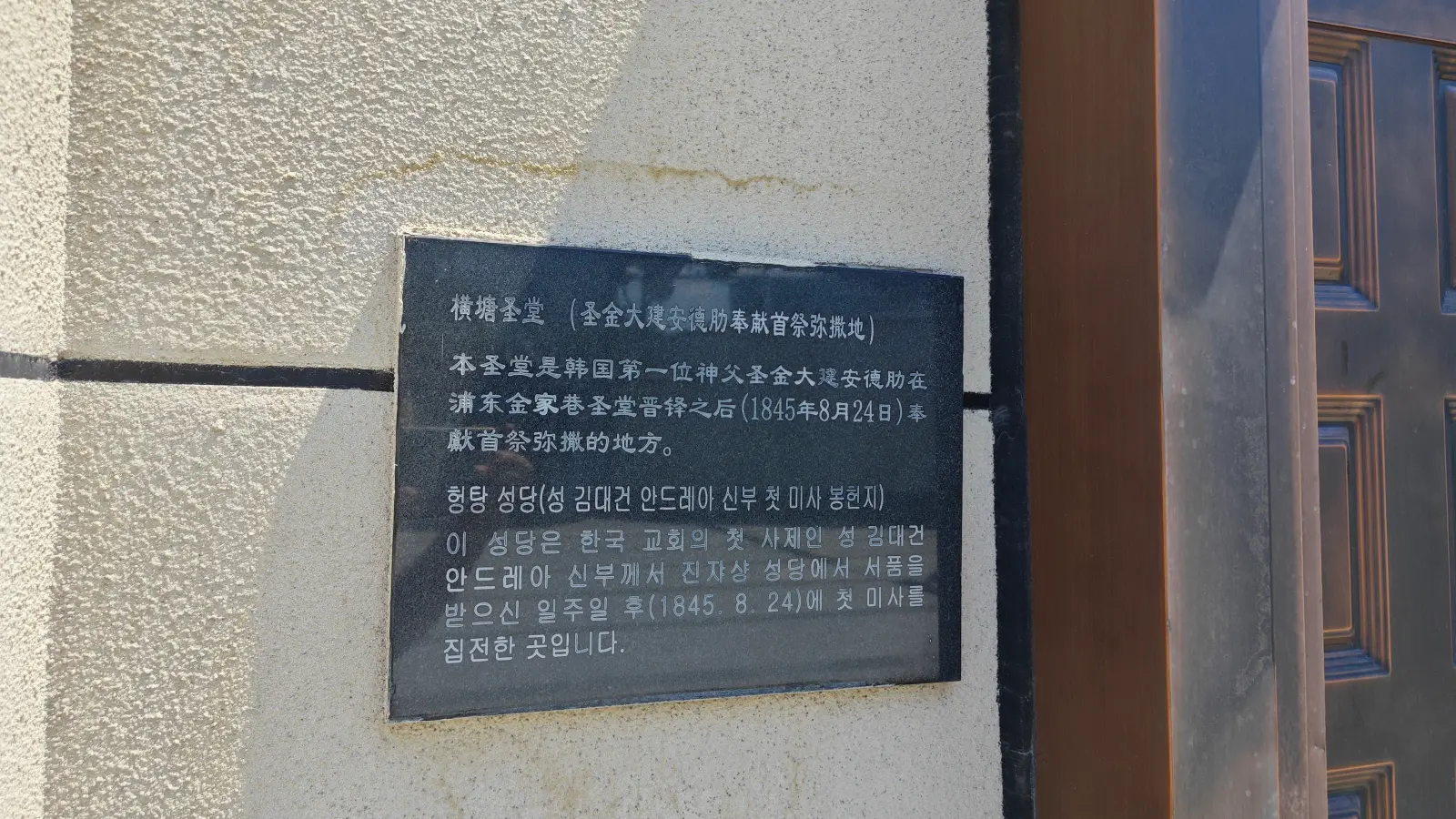 A dark plaque with Chinese and Korean text mounted on a cream wall next to a wooden door.